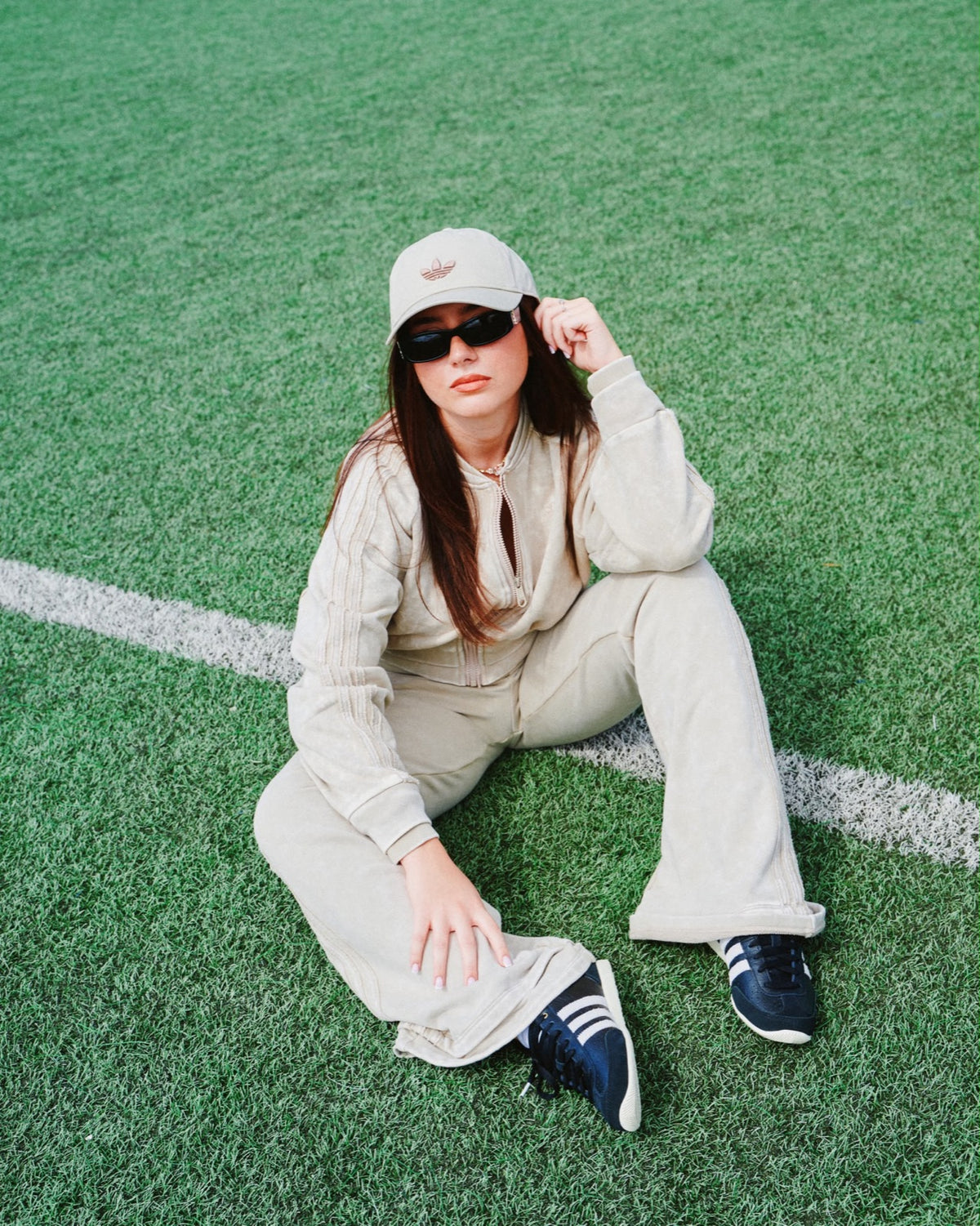 Ragazza in tuta beige con cappellino Adidas seduta sul campo